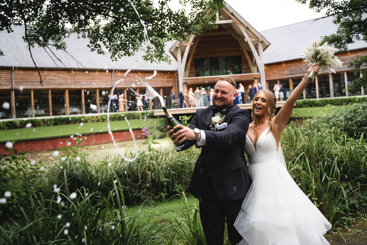 A couple shares a romantic moment outdoors at their wedding celebration, surrounded by lush greenery and rustic architecture.