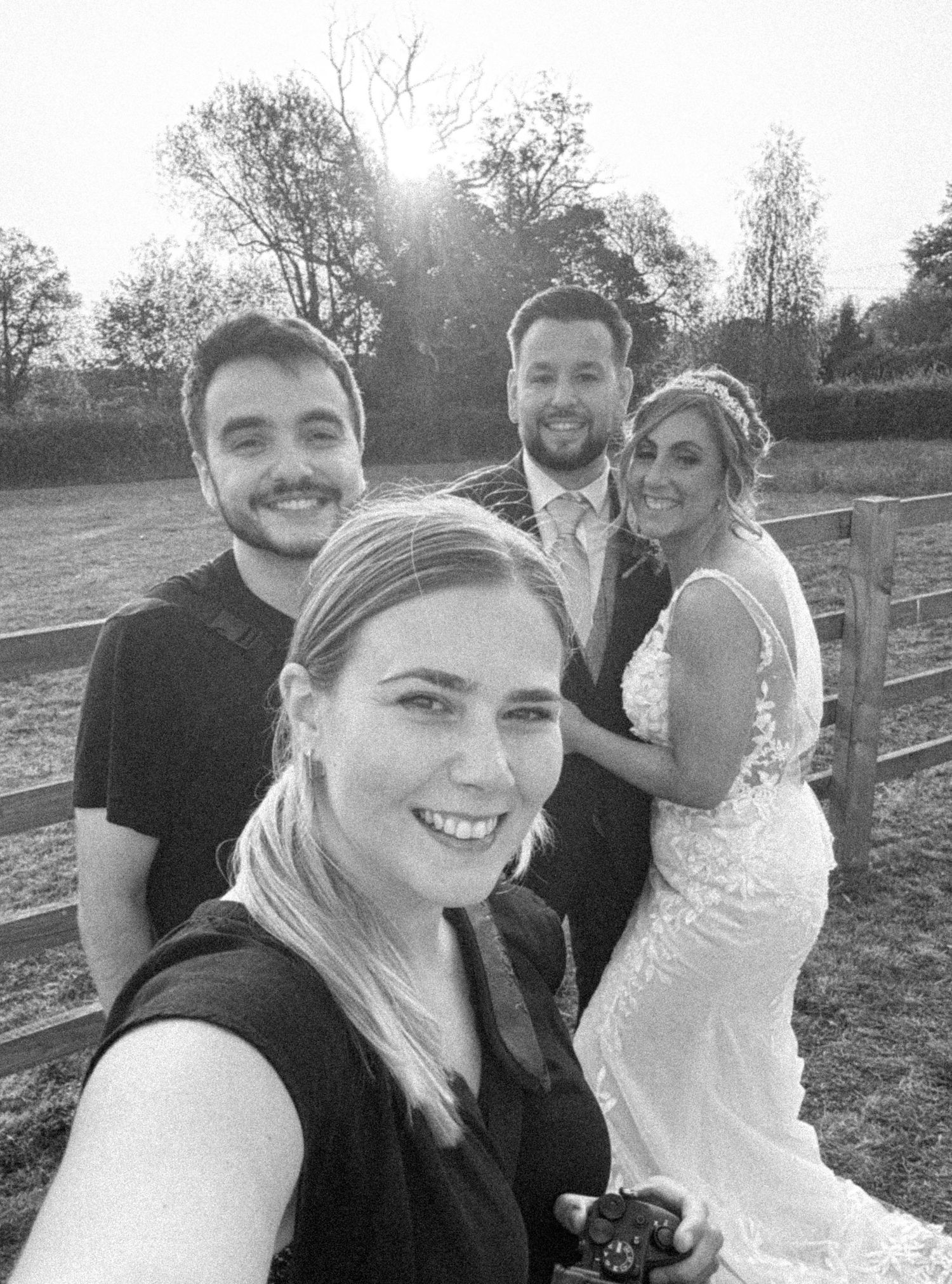 A black and white selfie photo taken outdoors at a wedding celebration with a rustic fence and trees in the background.