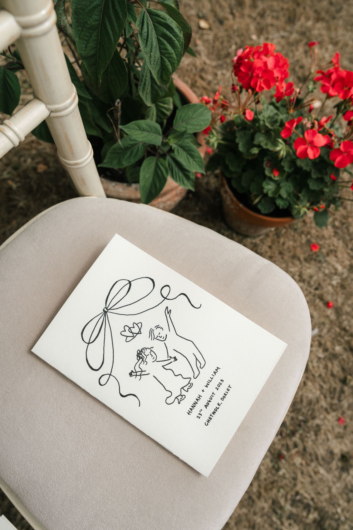 A white business card with minimalist line art illustration sits on a gray chair with red flowers in the background.
