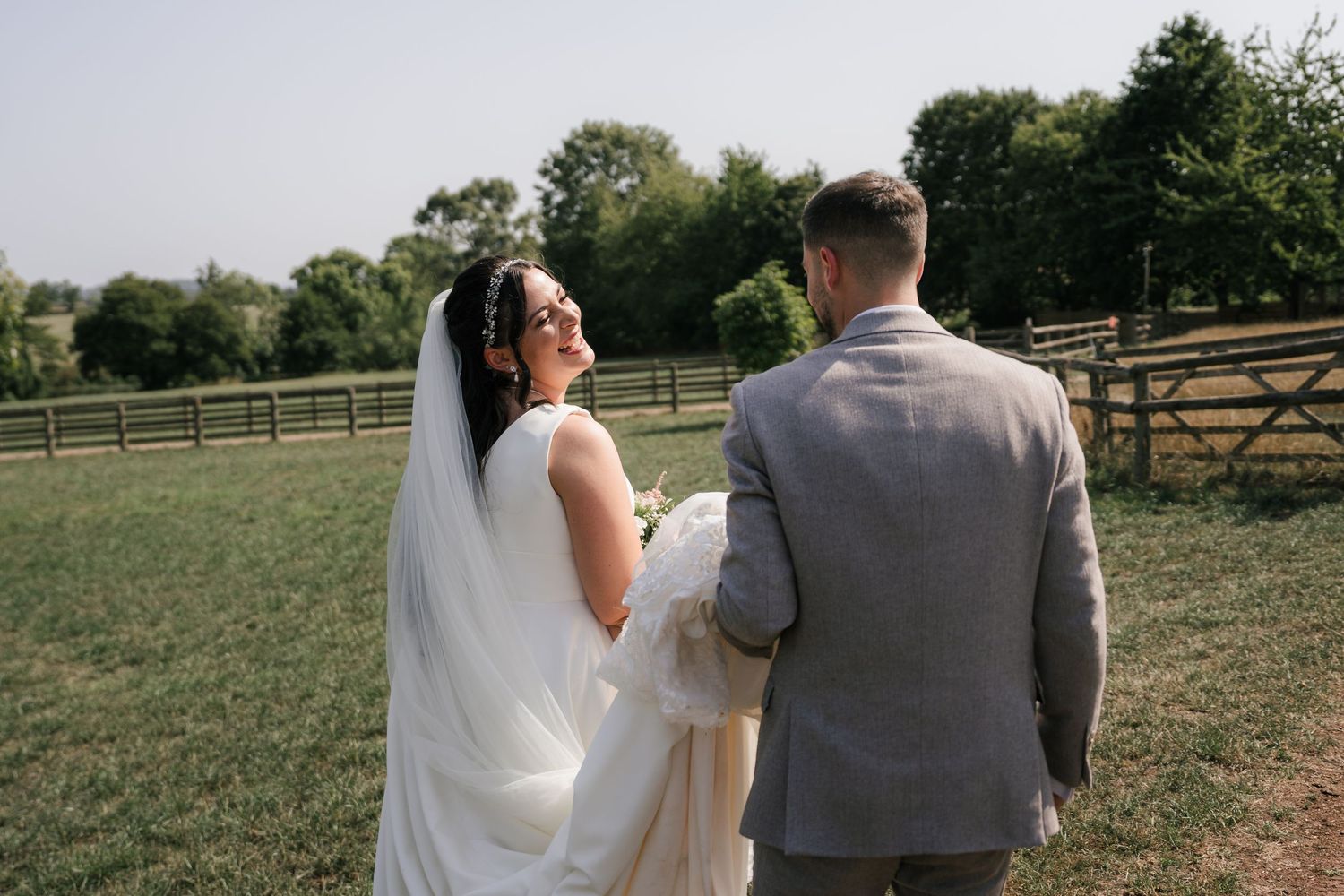 A couple shares a romantic moment in a rustic outdoor setting with wooden fences and green fields on their wedding day.