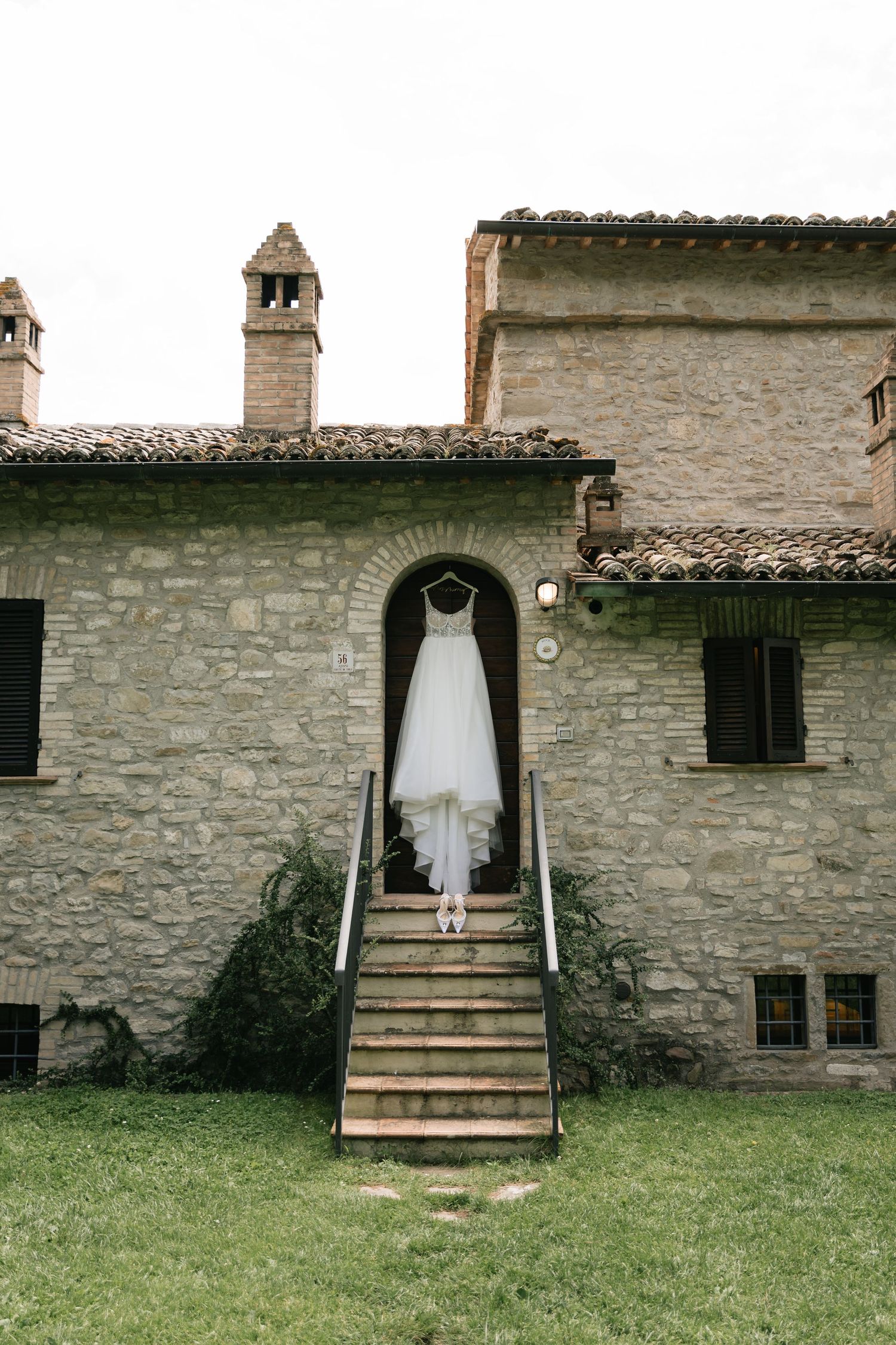 A white wedding dress displayed on stone stairs against a rustic stone wall exterior with grass in the foreground.
