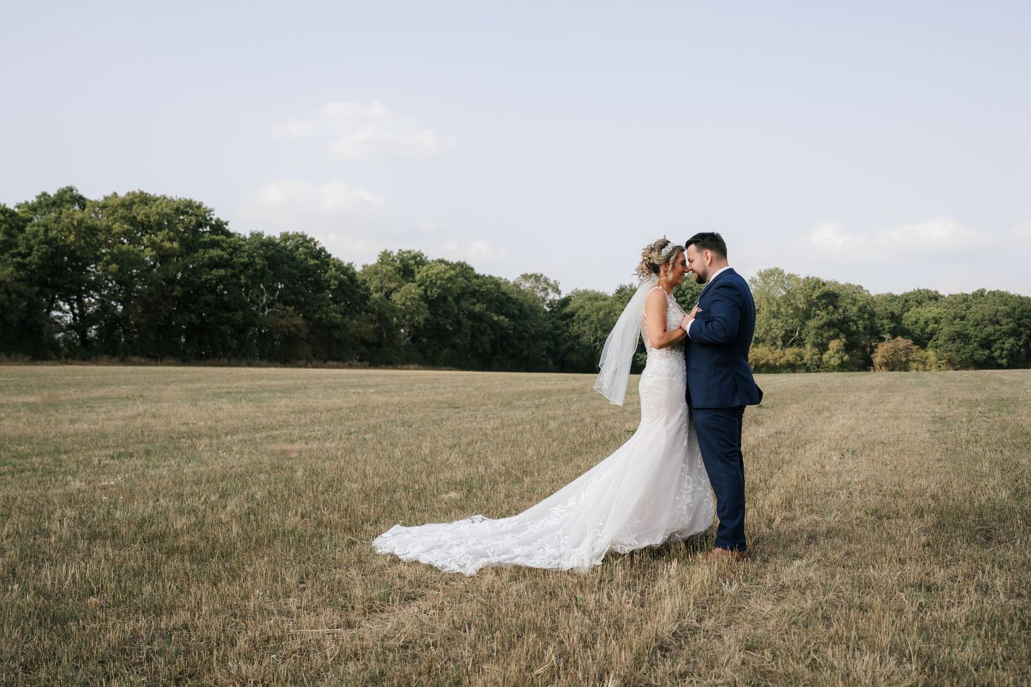 A couple in formal attire stands together in a golden grassy field on a sunny day.