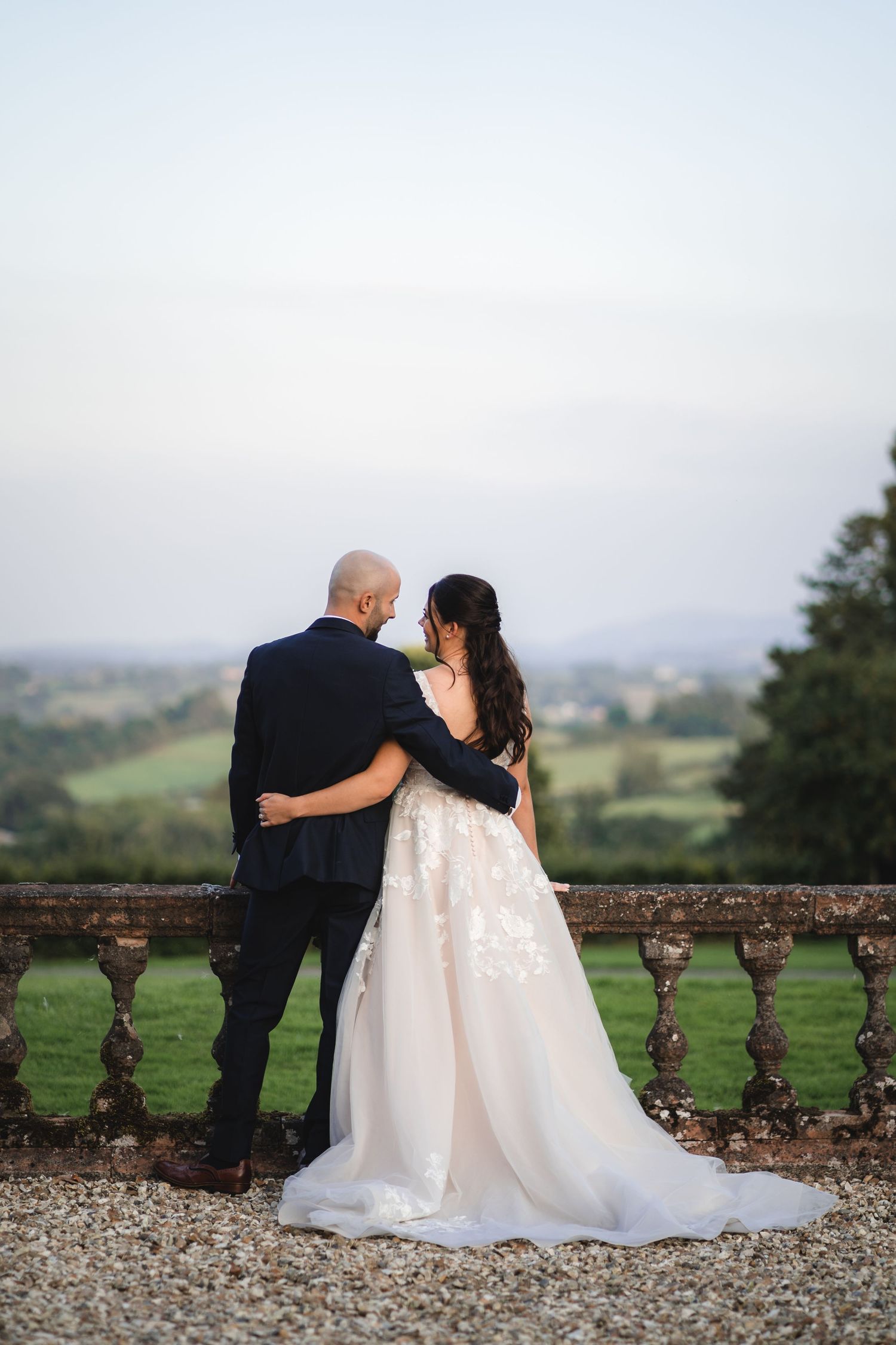 A couple embraces while looking out over a scenic countryside view from a stone balustrade at their wedding.
