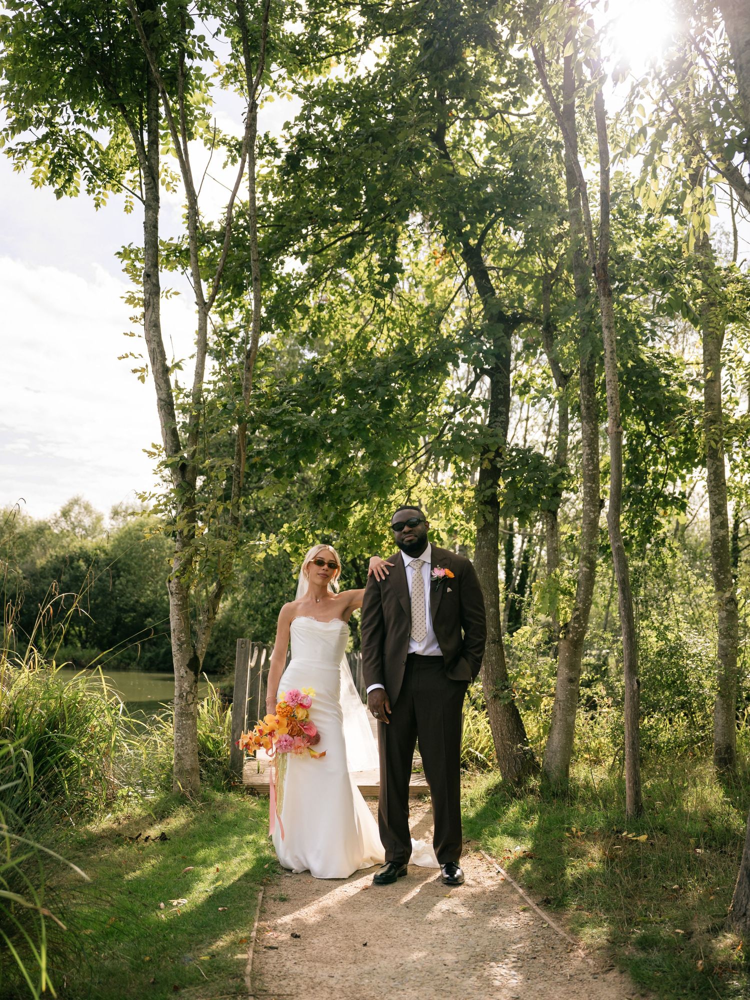 Bride and groom walking hand in hand on a sunlit garden path surrounded by lush green trees on their wedding day.