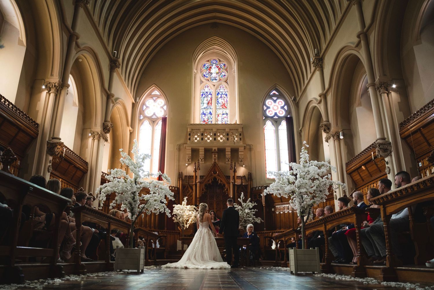 A panoramic view of a church interior with Gothic arches, stained glass windows, and wooden pews during a wedding ceremony.