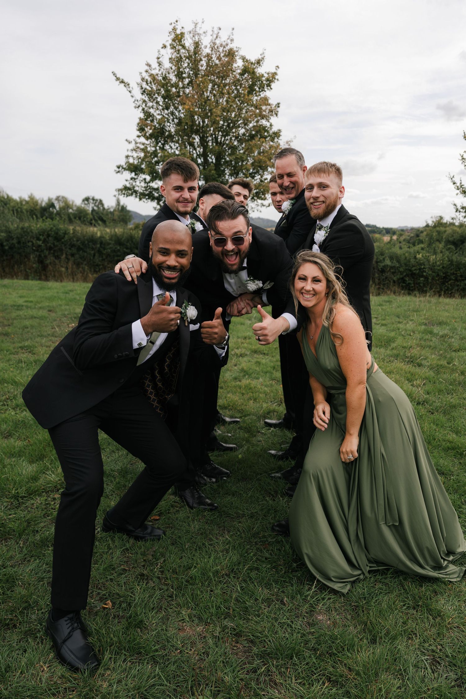 Wedding party members in black suits and olive green dresses pose playfully together on grassy field outdoors.