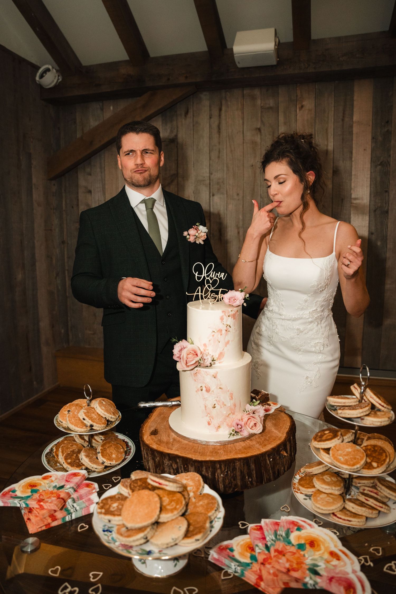 Wedding reception dessert table features a pink-accented white cake alongside stacks of cookies and pastries in a rustic barn setting.