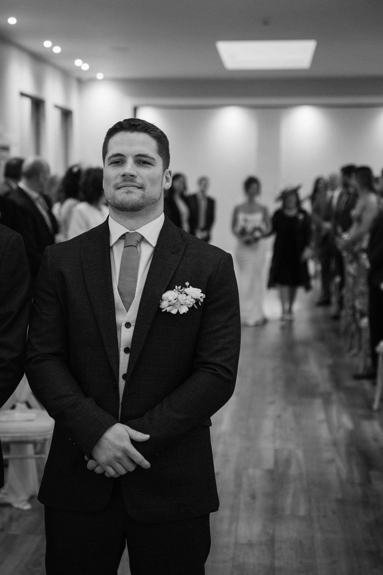 A wedding attendant in a dark suit and tie with a white boutonniere stands in a reception hall with guests in background.