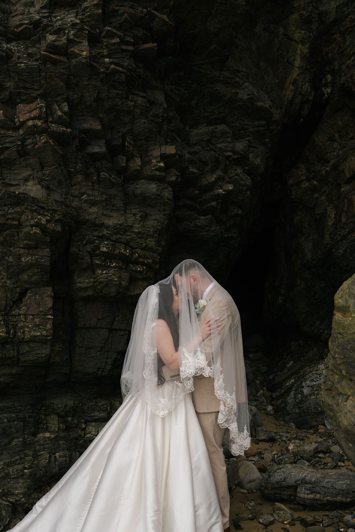 Bride and groom kissing under a lace veil against a dramatic dark rocky backdrop in a romantic wedding portrait.