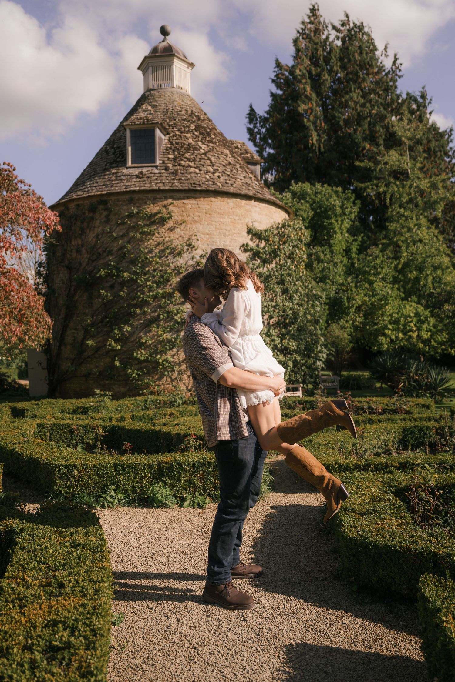 A romantic moment captured in a garden path with stone buildings and lush greenery in the background.