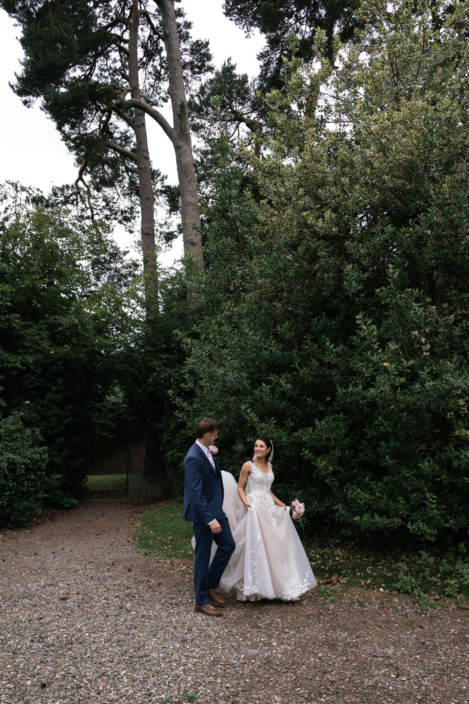 A couple in wedding attire stands together on a gravel path surrounded by lush green trees in a forest setting.