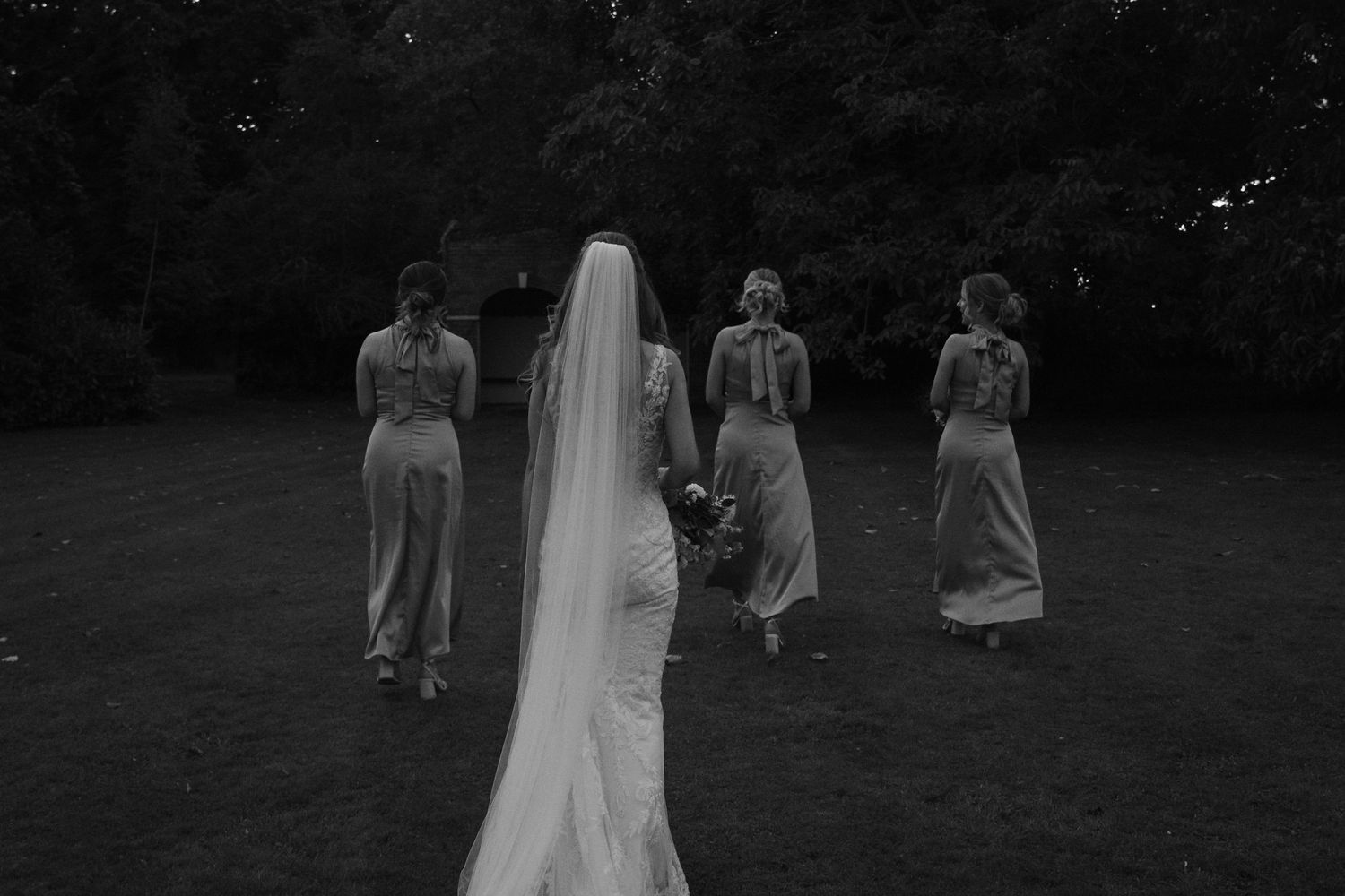 A group of people walk through a grassy outdoor area during a formal event with someone wearing a flowing white dress.