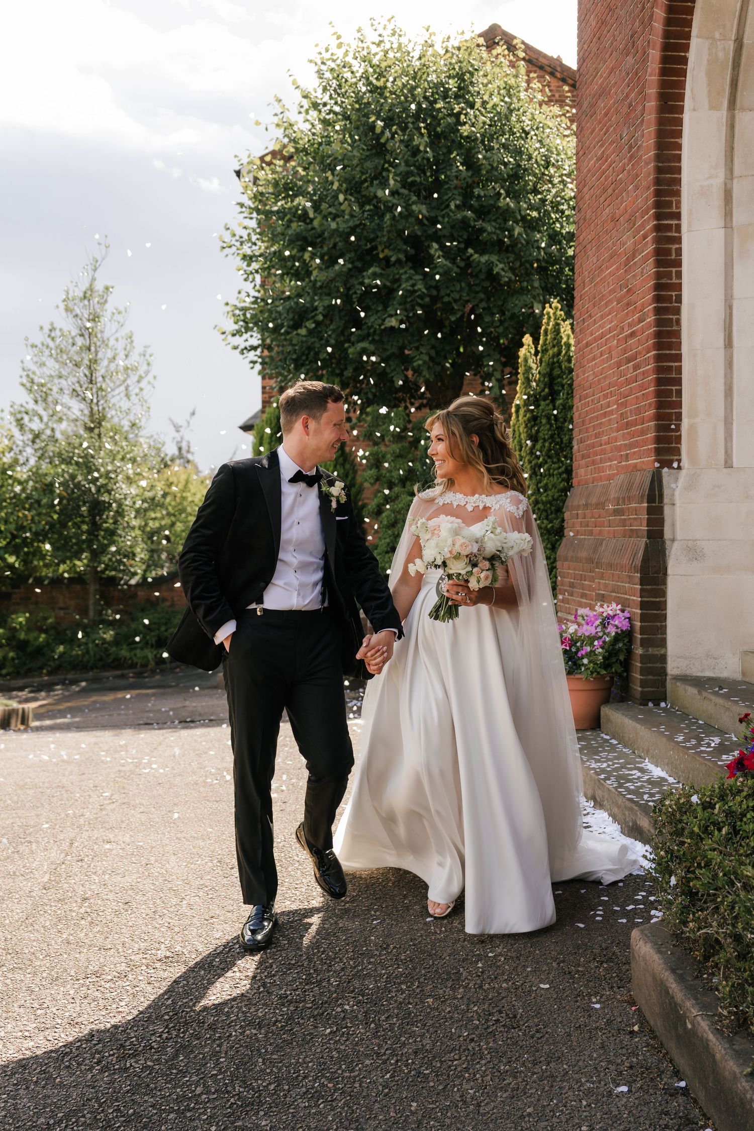 A couple in wedding attire walk hand in hand along a path beside a brick building on their special day.
