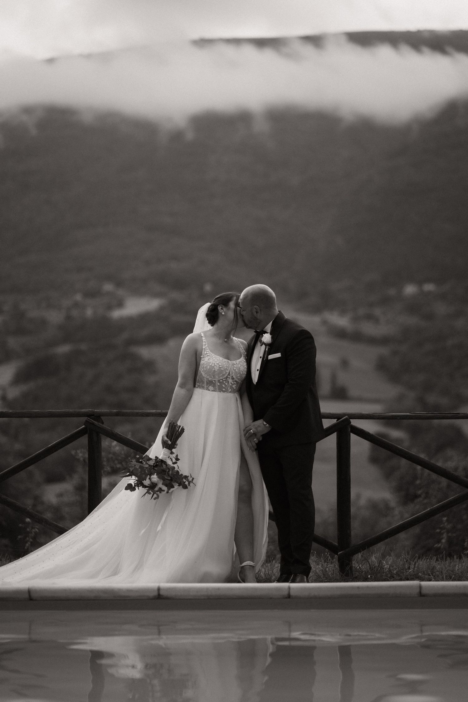 A romantic black and white wedding photo shows a couple embracing on a balcony overlooking misty mountains.