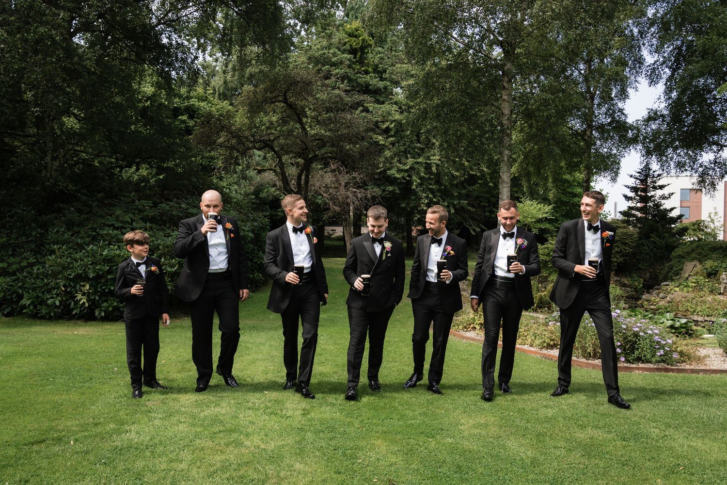 Groomsmen in black suits stand in a row on a lush green lawn during an outdoor wedding ceremony.