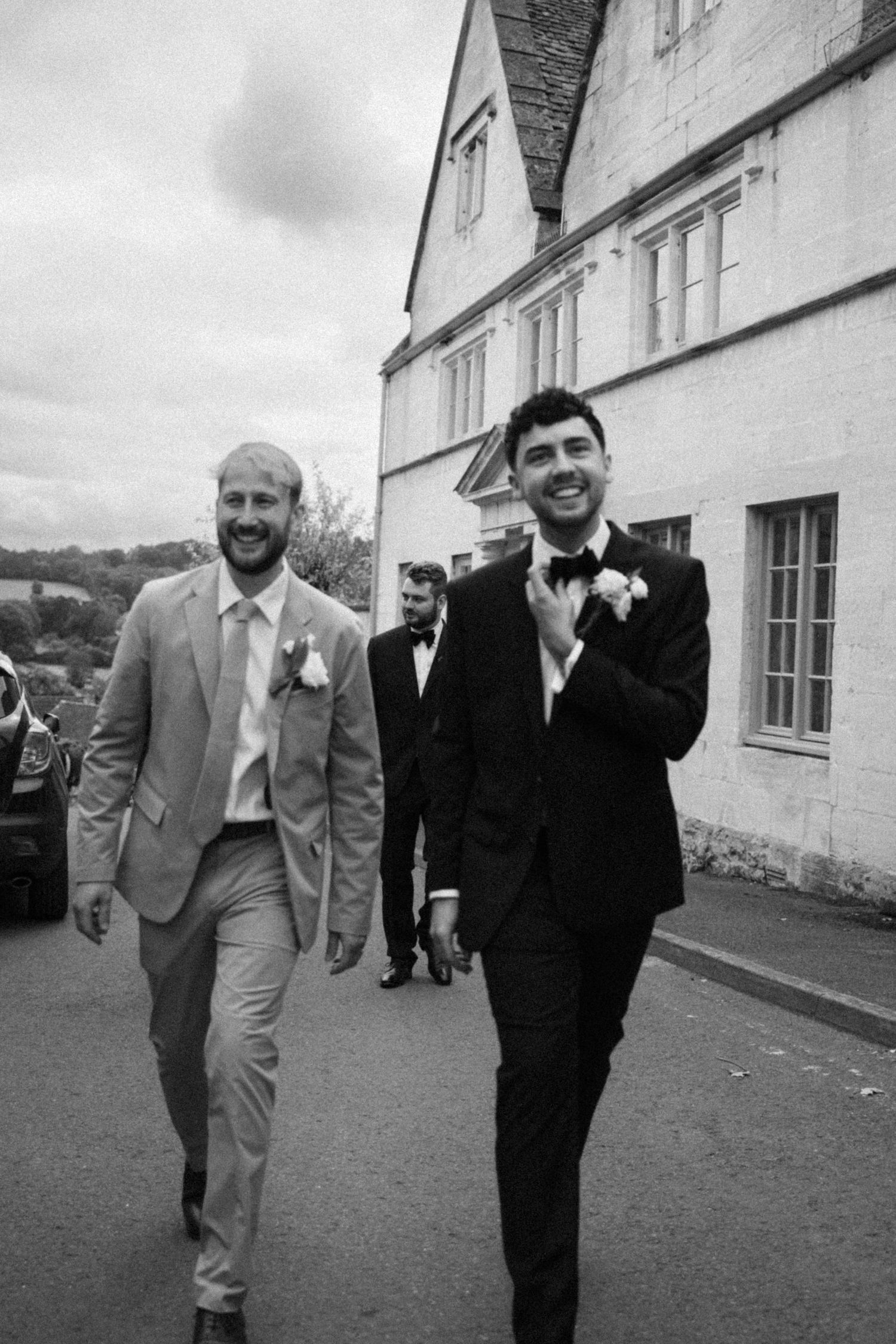 A series of black and white photos showing wedding party members in suits walking along a white building exterior.