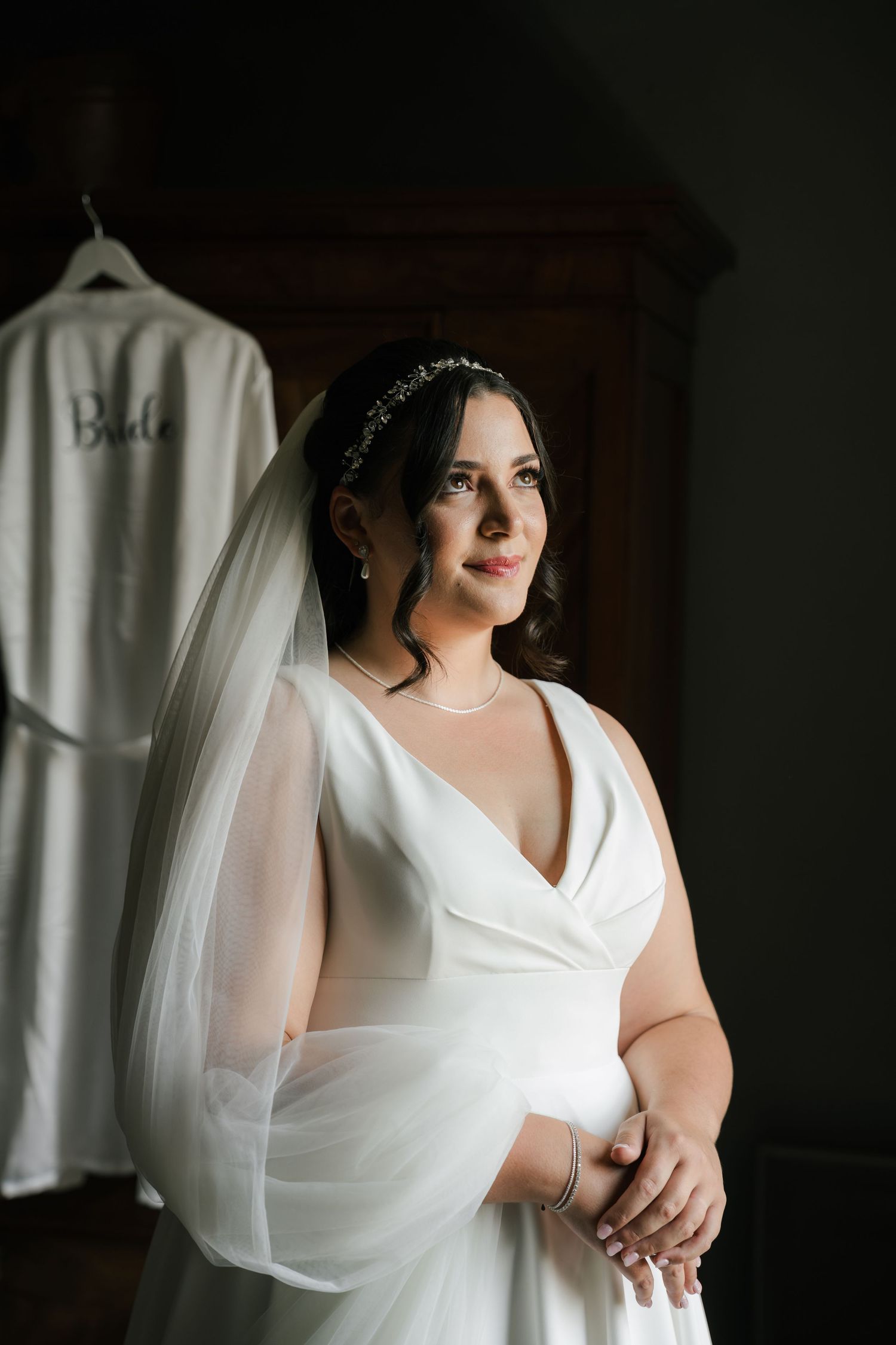 A bride in a classic white wedding dress poses thoughtfully in soft window light with flowing veil details.