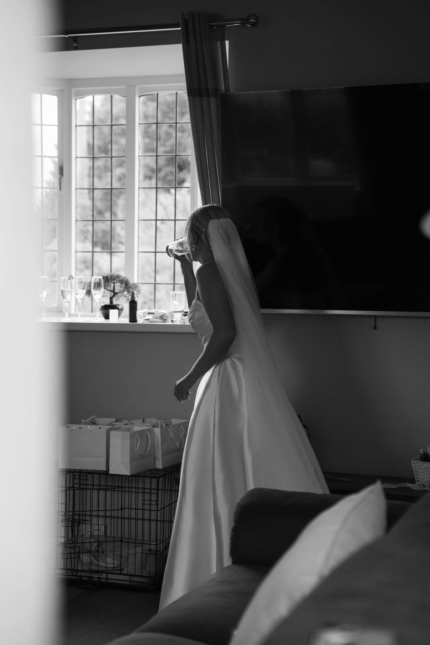 Black and white photograph of someone in a white dress standing by a window in a minimalist interior setting.