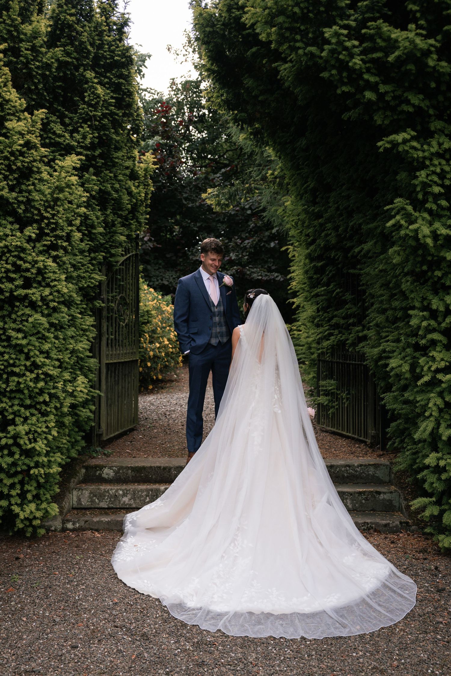 A romantic wedding couple walks along a garden path surrounded by lush greenery and iron gates.