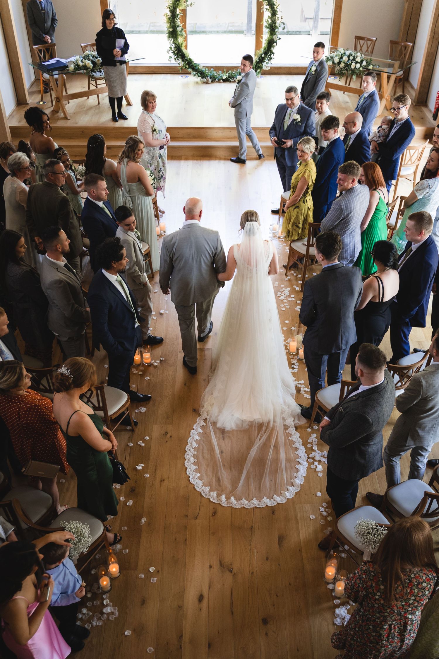 Wedding ceremony guests stand and watch as the bride walks down a rustic wooden aisle in an indoor venue.