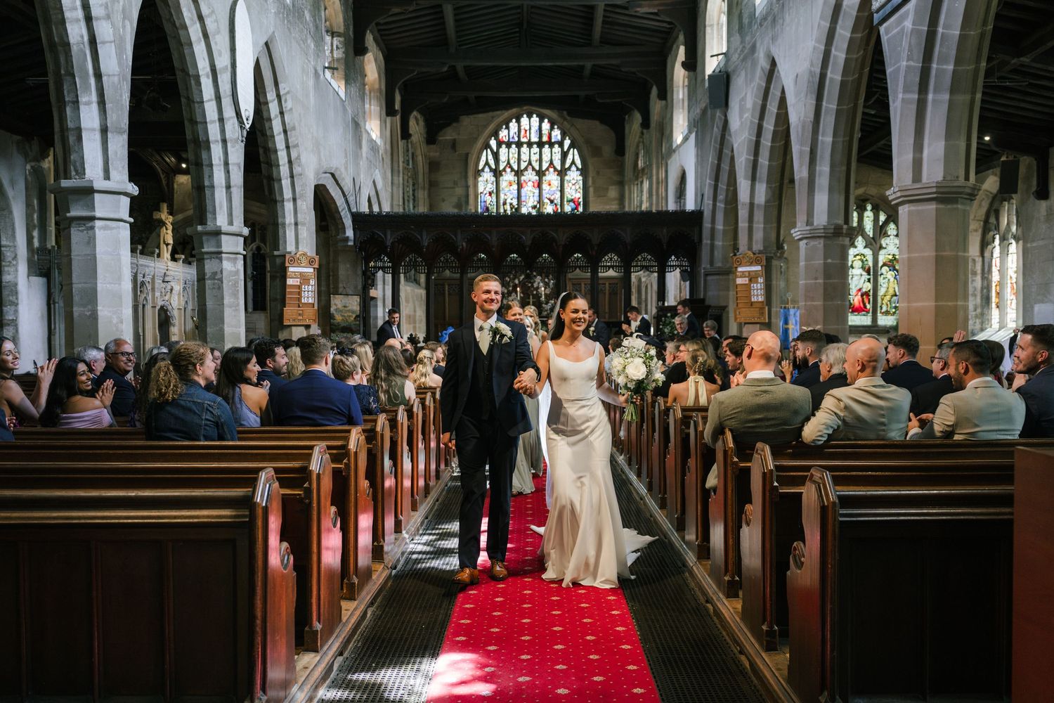 Bride and groom walk down red carpet aisle in historic stone church surrounded by wedding guests.