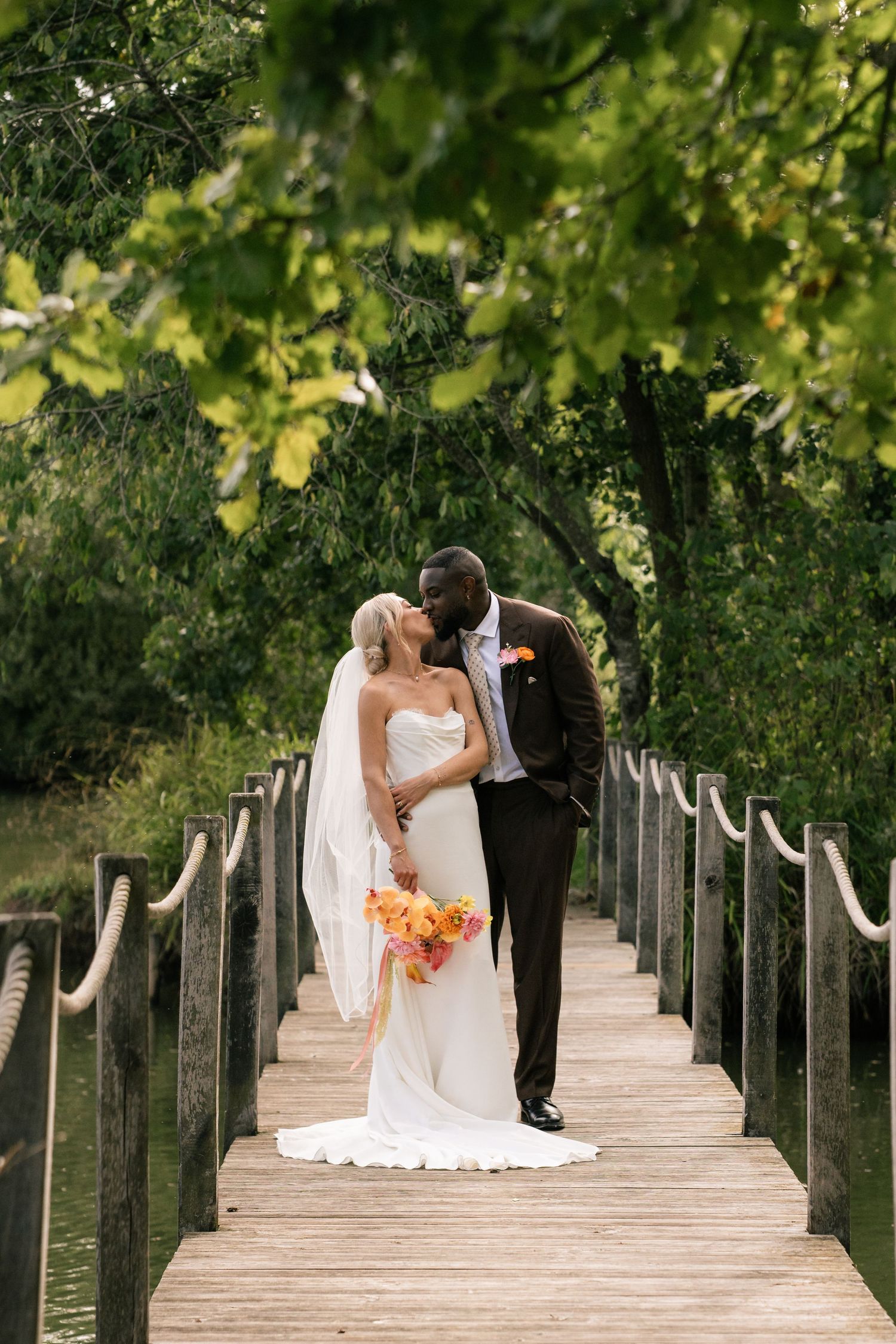 Interracial couple sharing a kiss on a wooden bridge surrounded by lush greenery, bride holding colorful bouquet.