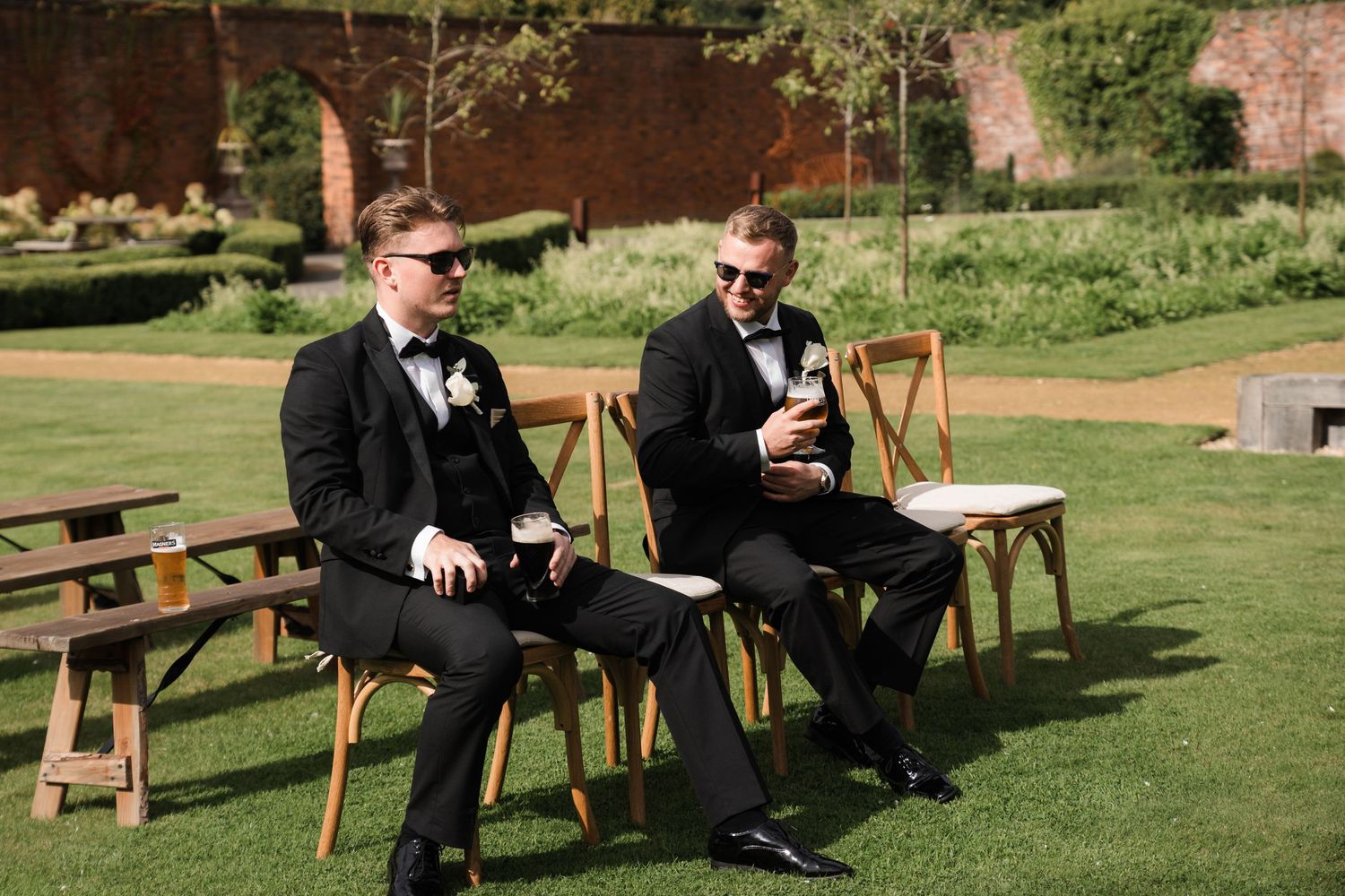 People in formal attire relax on wooden benches in a sunny outdoor garden setting.