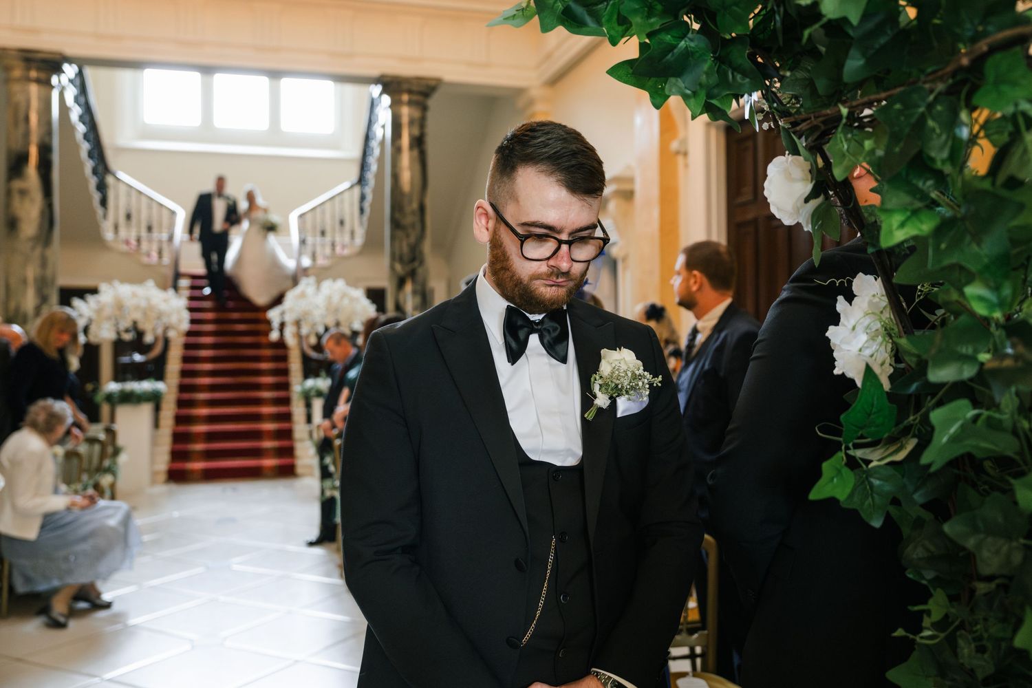 A wedding couple walks down an elegant staircase in a grand venue with natural light streaming through windows.