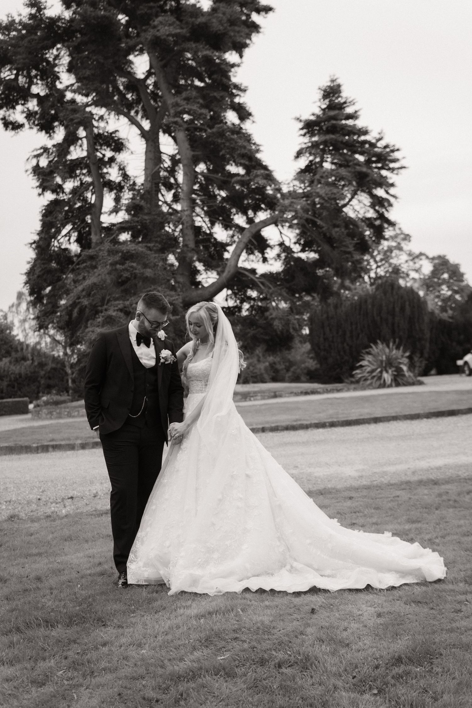 A black and white wedding photo showing a couple in formal attire standing on a lawn with tall trees in the background.