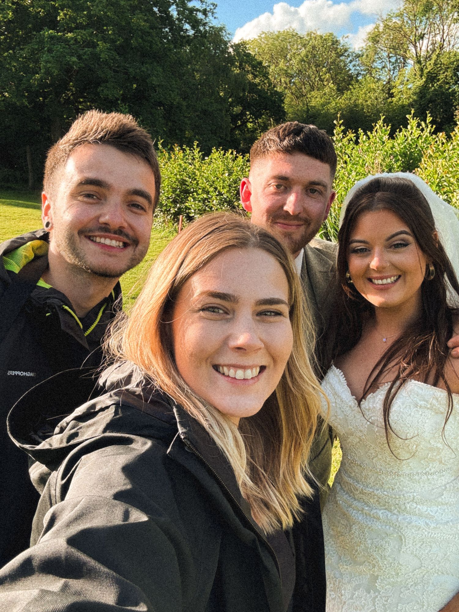 A group of friends smiling and taking selfies outdoors on a sunny day with green foliage in the background.
