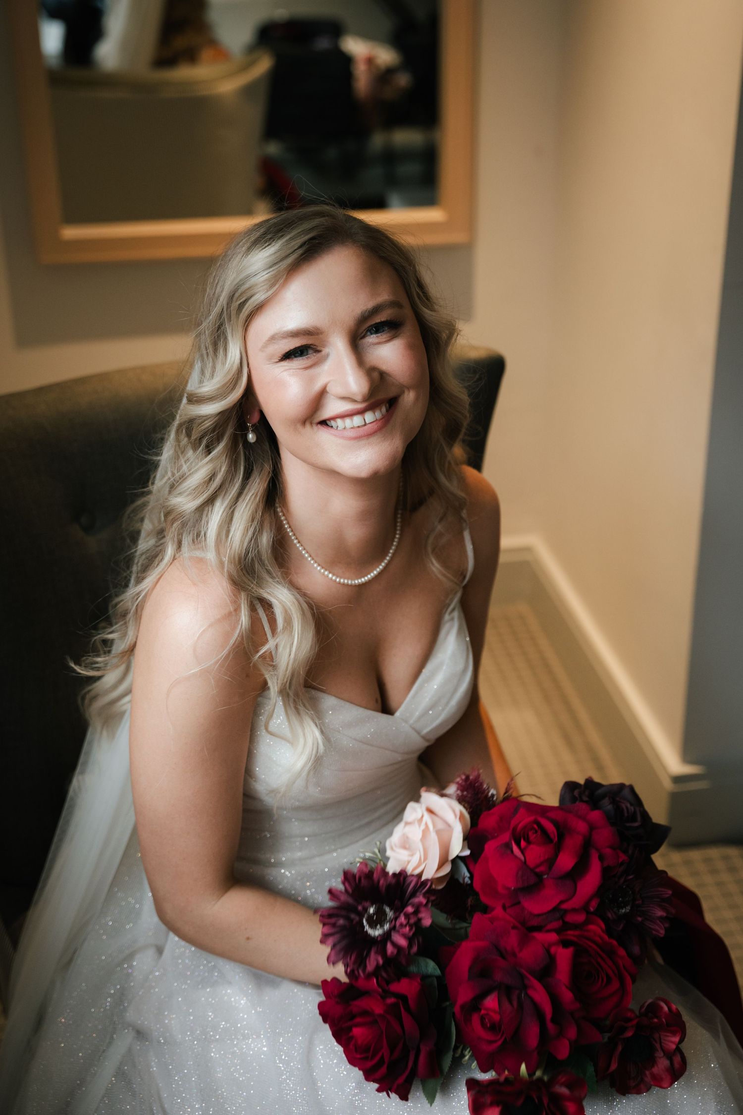 Bride in white wedding dress poses with deep red and burgundy rose bouquet in indoor setting.