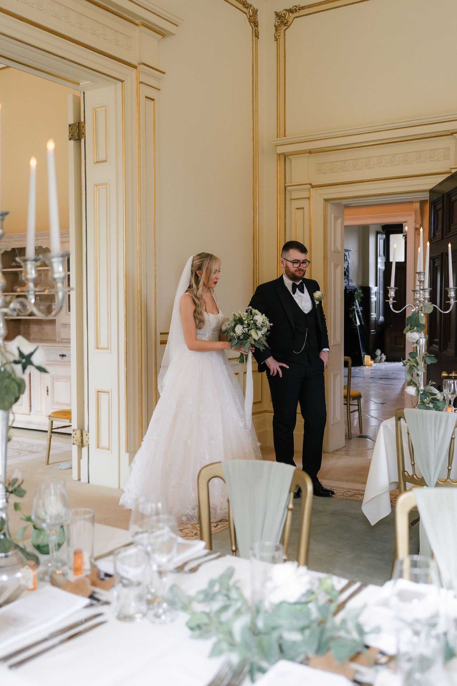 Elegant wedding reception setup in a cream-colored room with white candles and eucalyptus decor on dining tables.