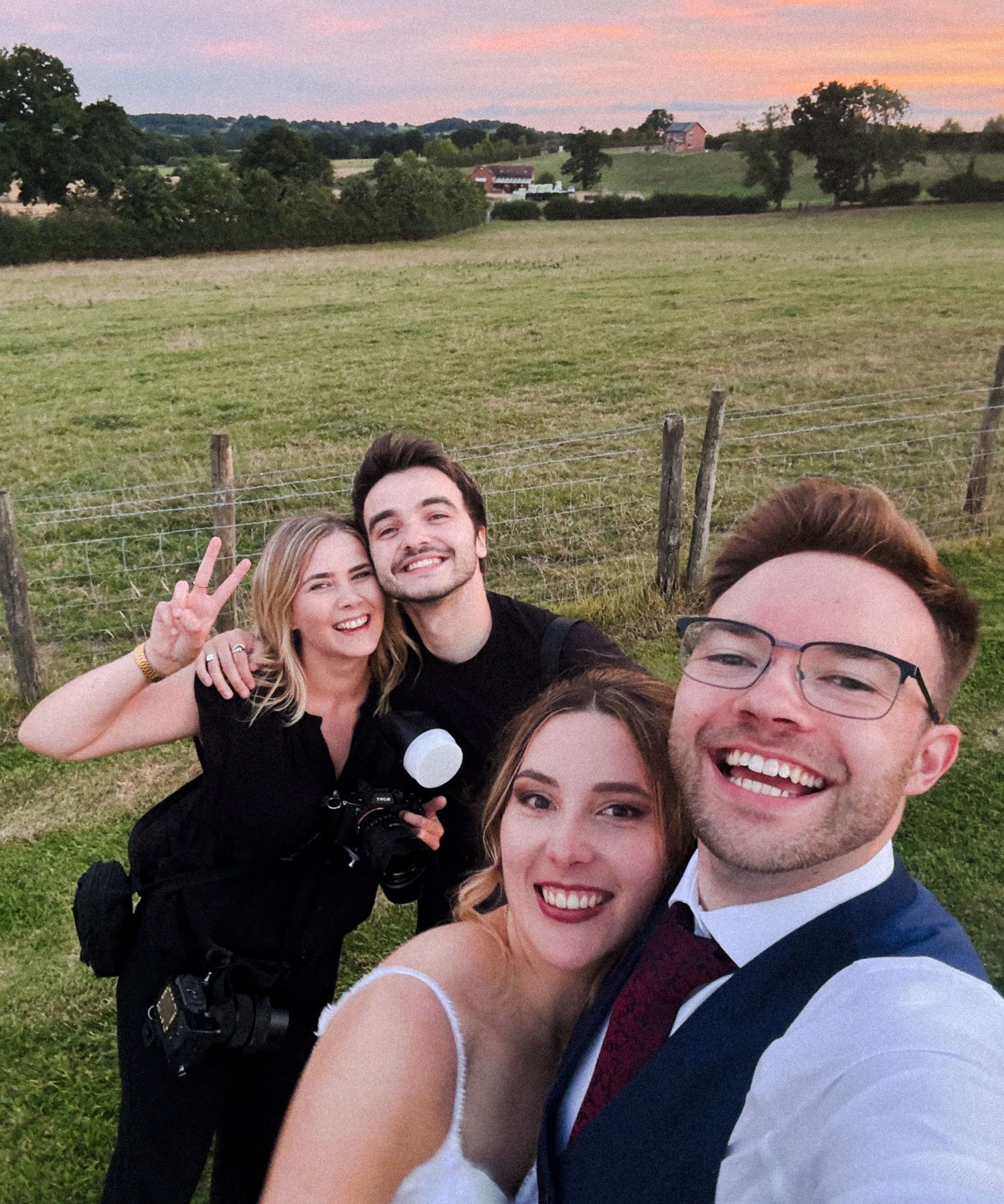 A couple takes selfies together in an outdoor field at sunset showing joyful expressions and peace signs.