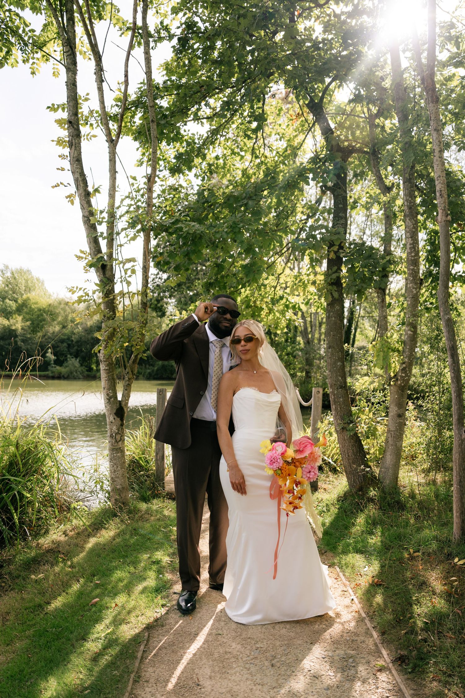 Bride in white gown with colorful bouquet and groom in brown suit pose on garden path by lake in sunny outdoor wedding.