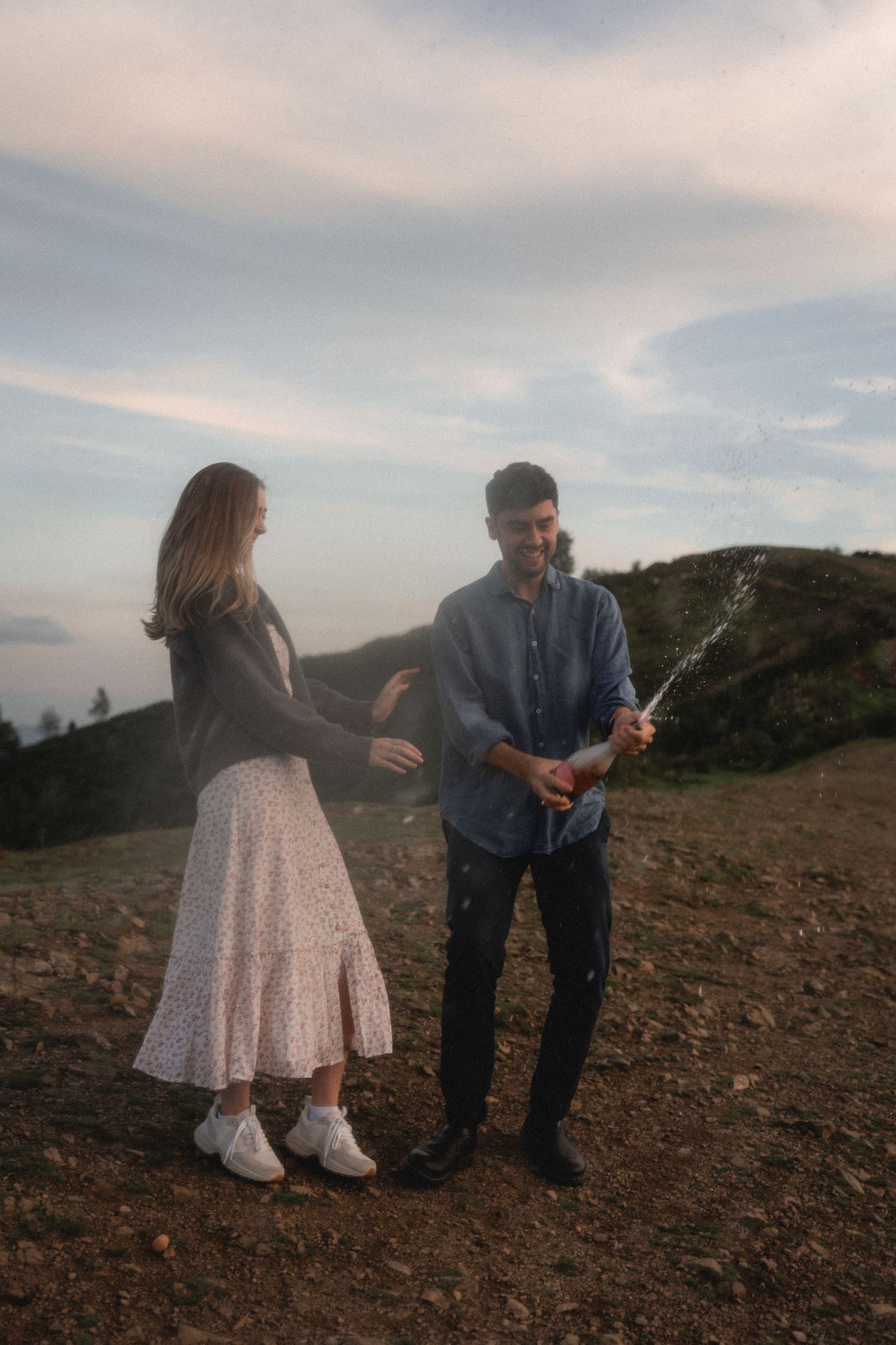 A romantic couple shares champagne together on a rocky hillside during sunset.