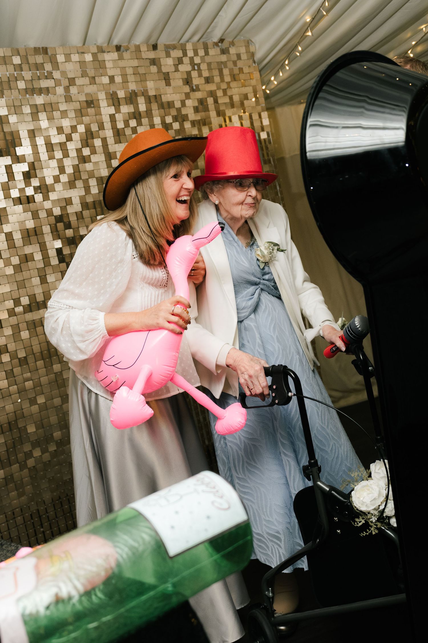 Two people in hats laughing and posing with a pink flamingo prop against a metallic mosaic wall at an event.