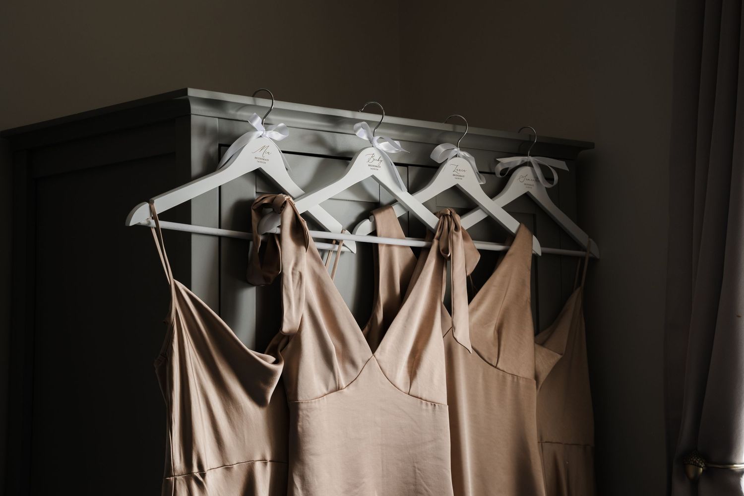 A row of beige bridesmaid dresses hanging on white hangers against a dark wall.