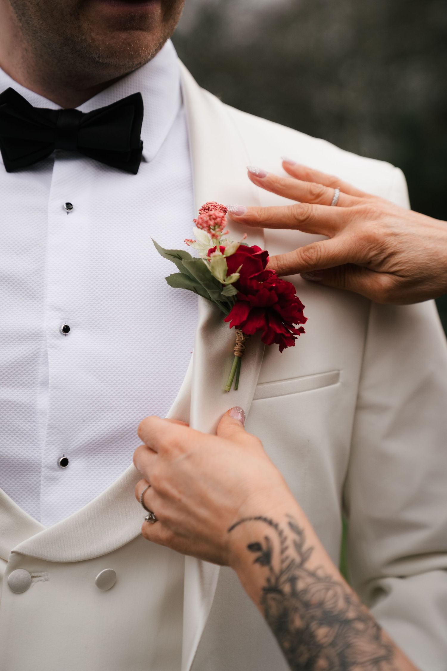 Hands with floral tattoos pin a deep red boutonniere onto a white tuxedo jacket.