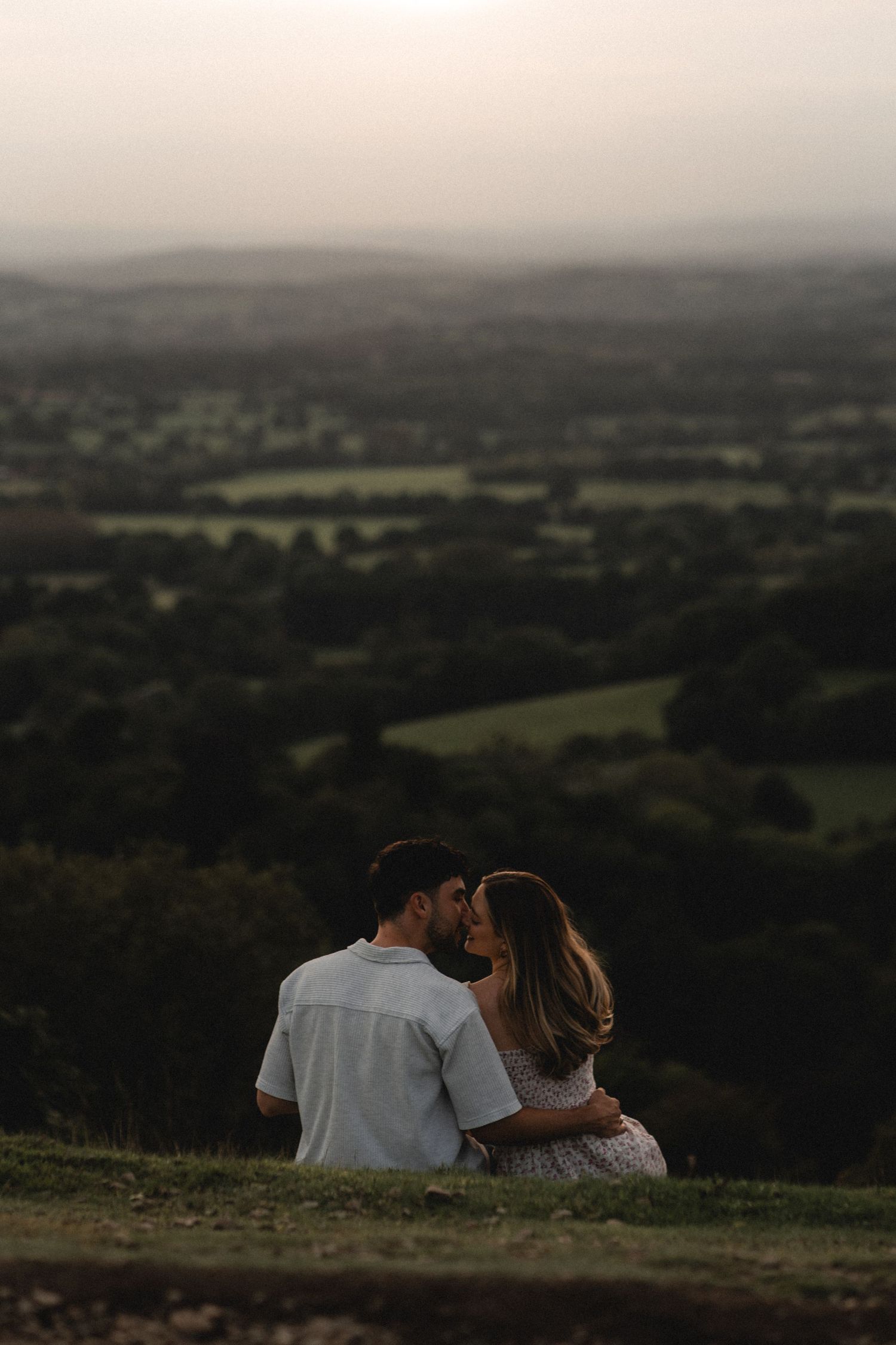 Couple sits together on a hilltop overlooking a vast countryside landscape at dusk.