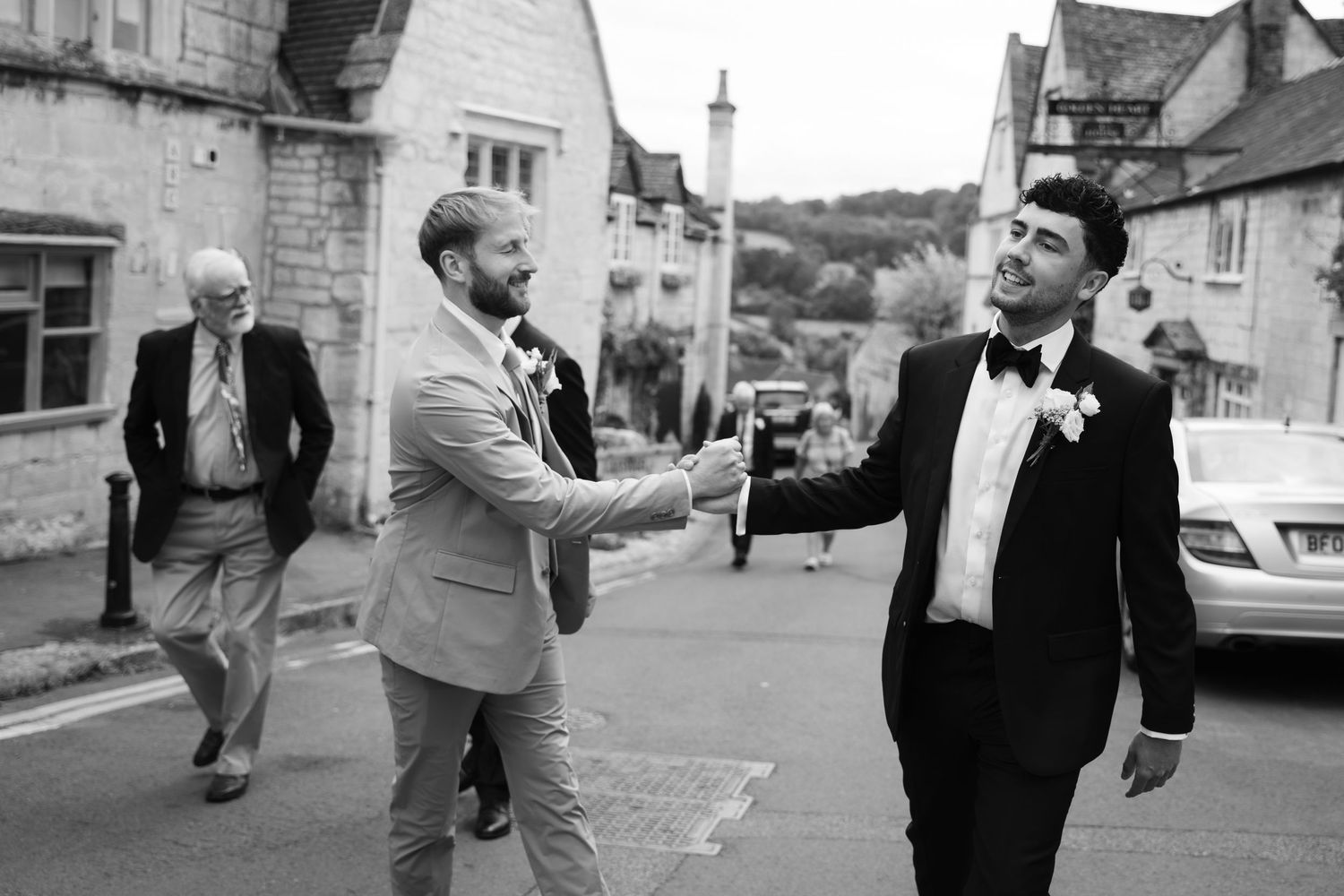 Two people in formal attire shake hands on a quaint village street while another person walks in the background.