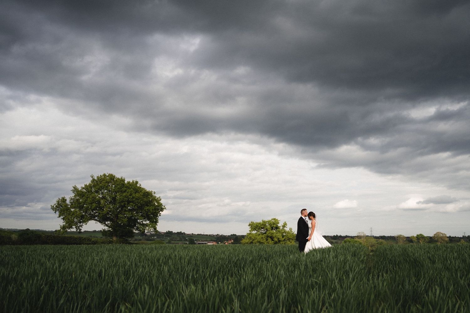 A dramatic panoramic view of stormy clouds rolling over a green wheat field with a white farmhouse in the distance.