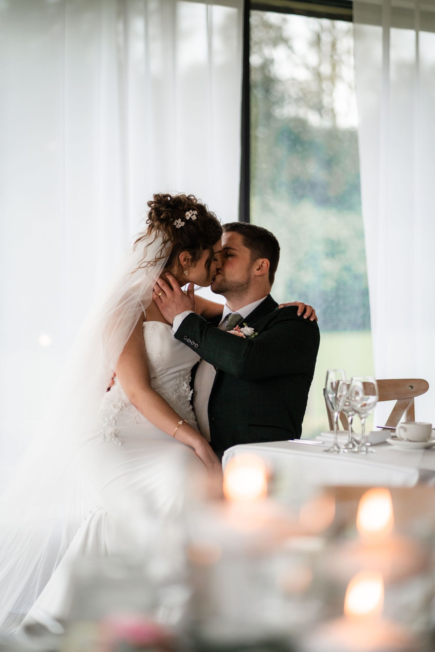 Romantic wedding couple shares intimate kiss at reception table with soft candlelight and white decor.