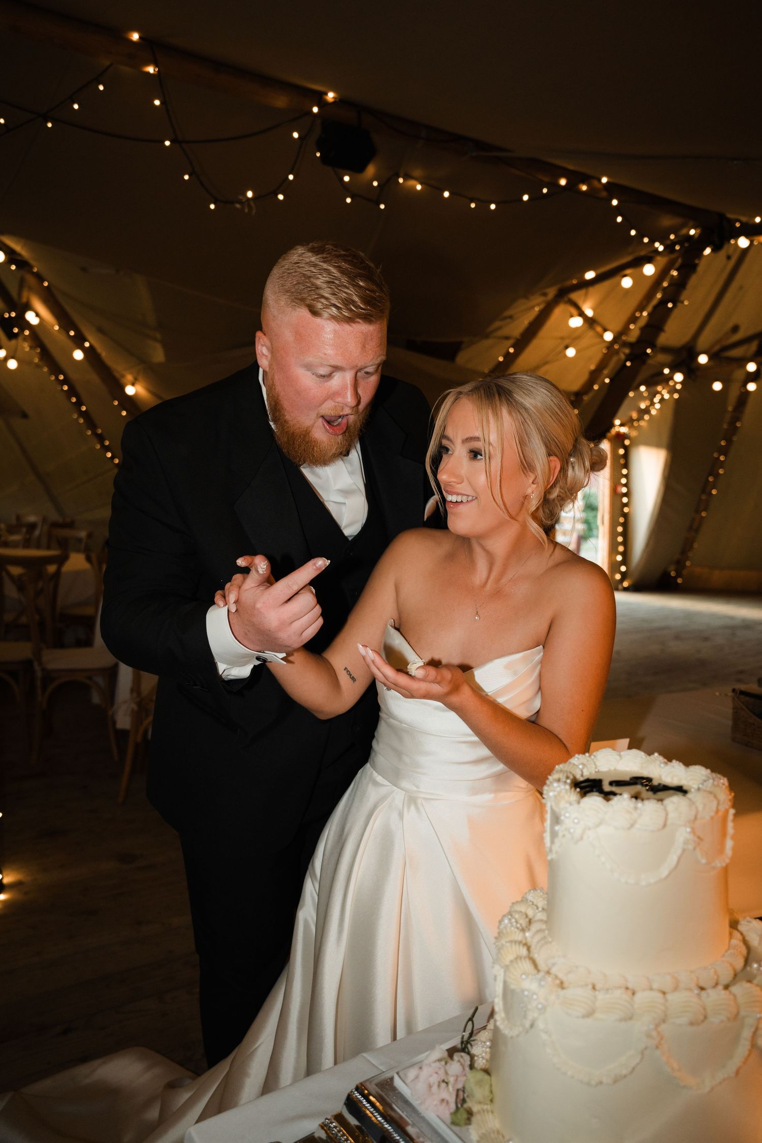 Newlywed couple shares playful moment while cutting their white wedding cake together at a reception under string lights.