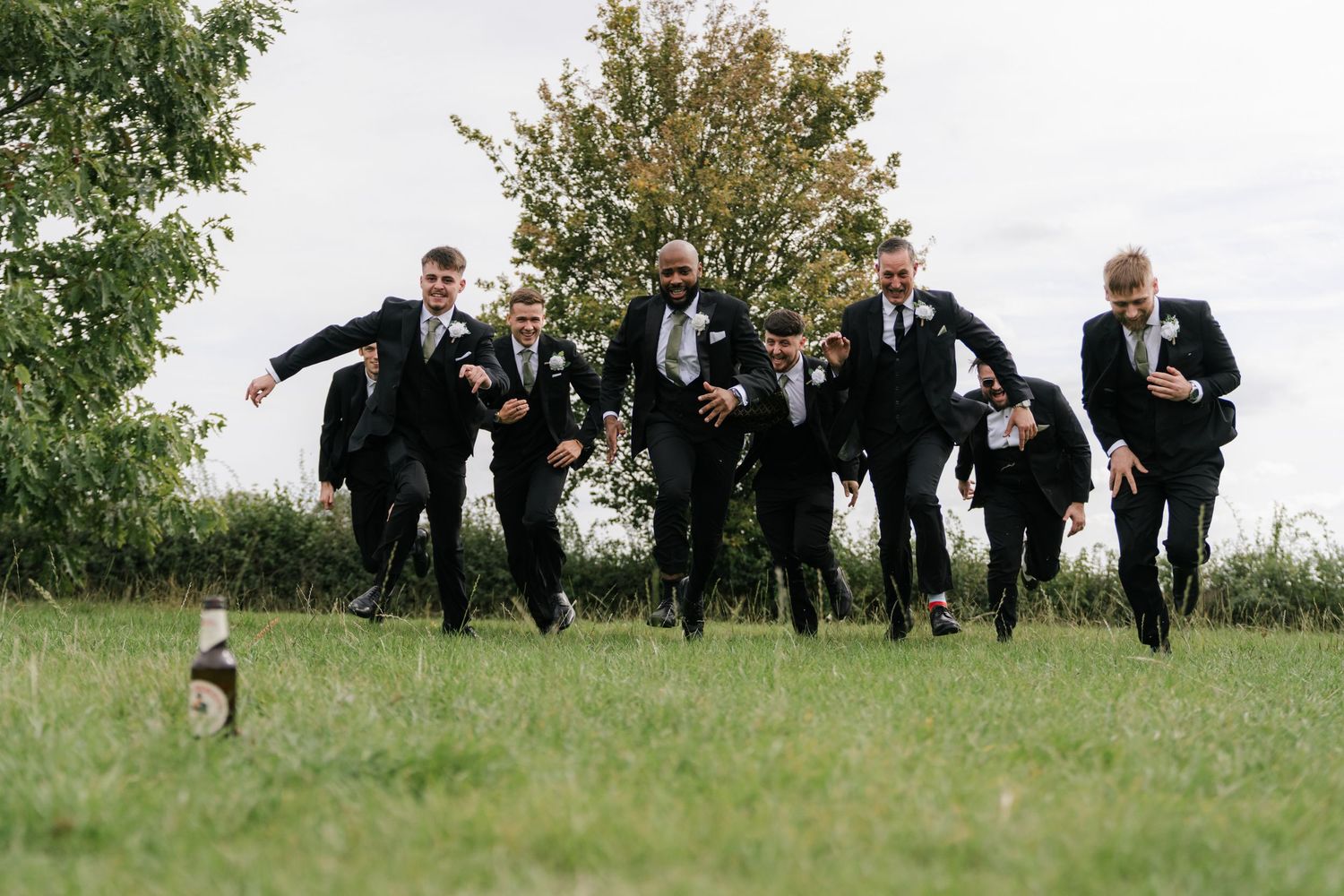 A group in formal black suits playfully runs through a grassy field during a wedding photoshoot.