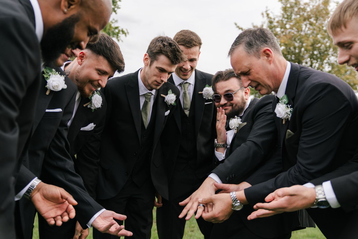 Groomsmen in black suits huddle together laughing while looking down at something in their hands outdoors.