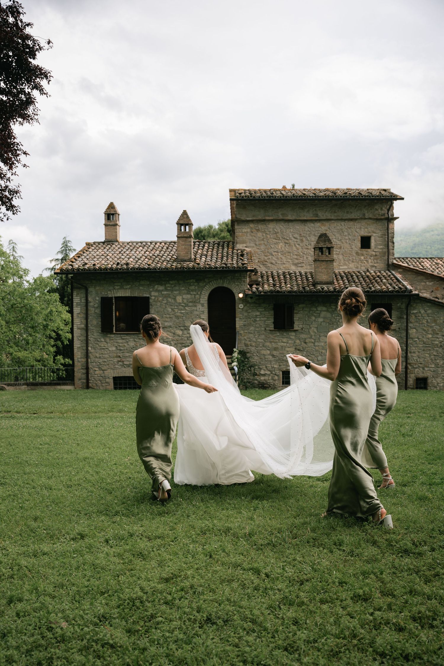 Wedding party carries flowing white dress across green field with rustic stone buildings in background.