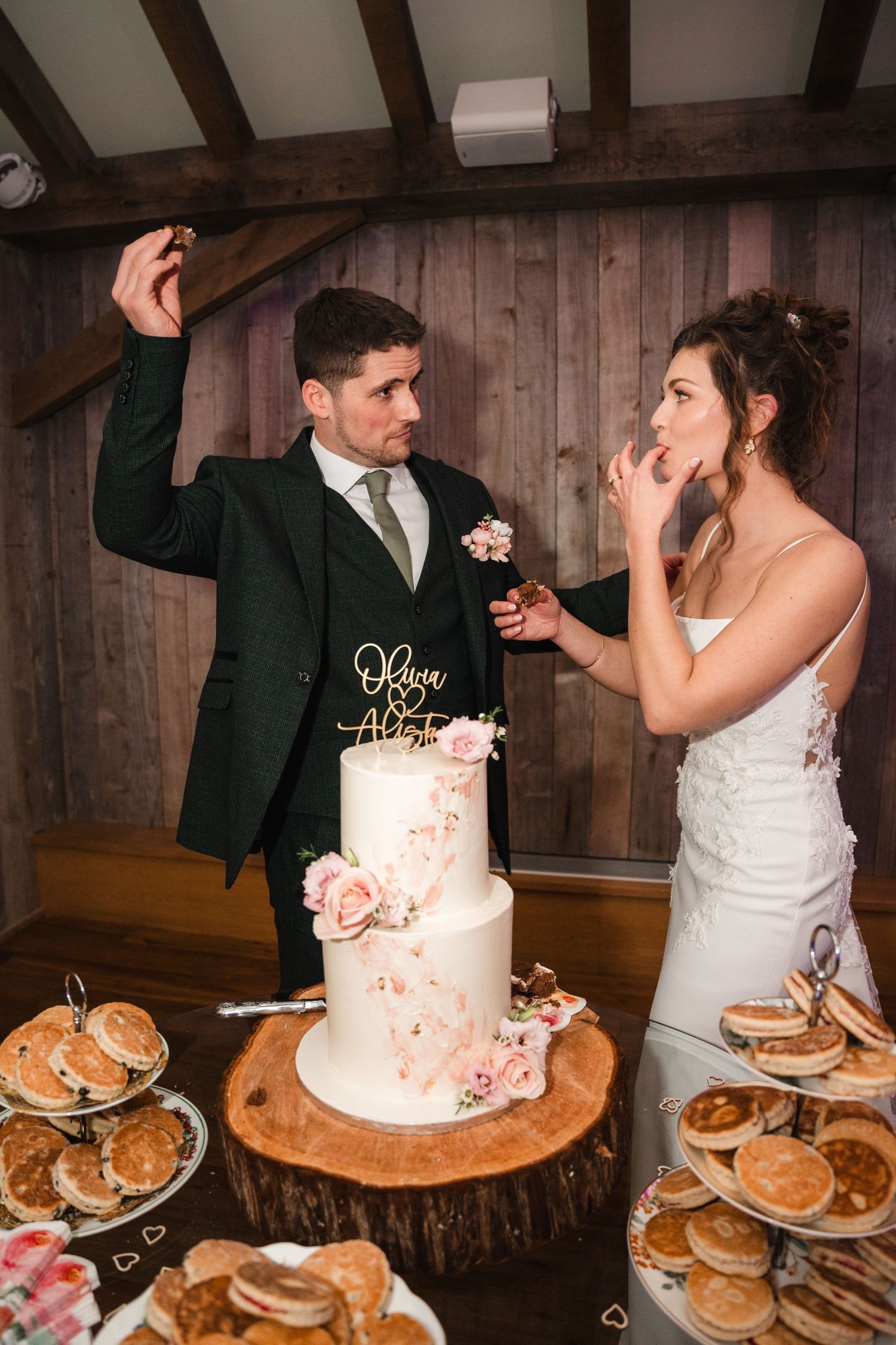 Wedding couple shares a playful moment while cutting their rustic wedding cake surrounded by donuts on wooden stands.