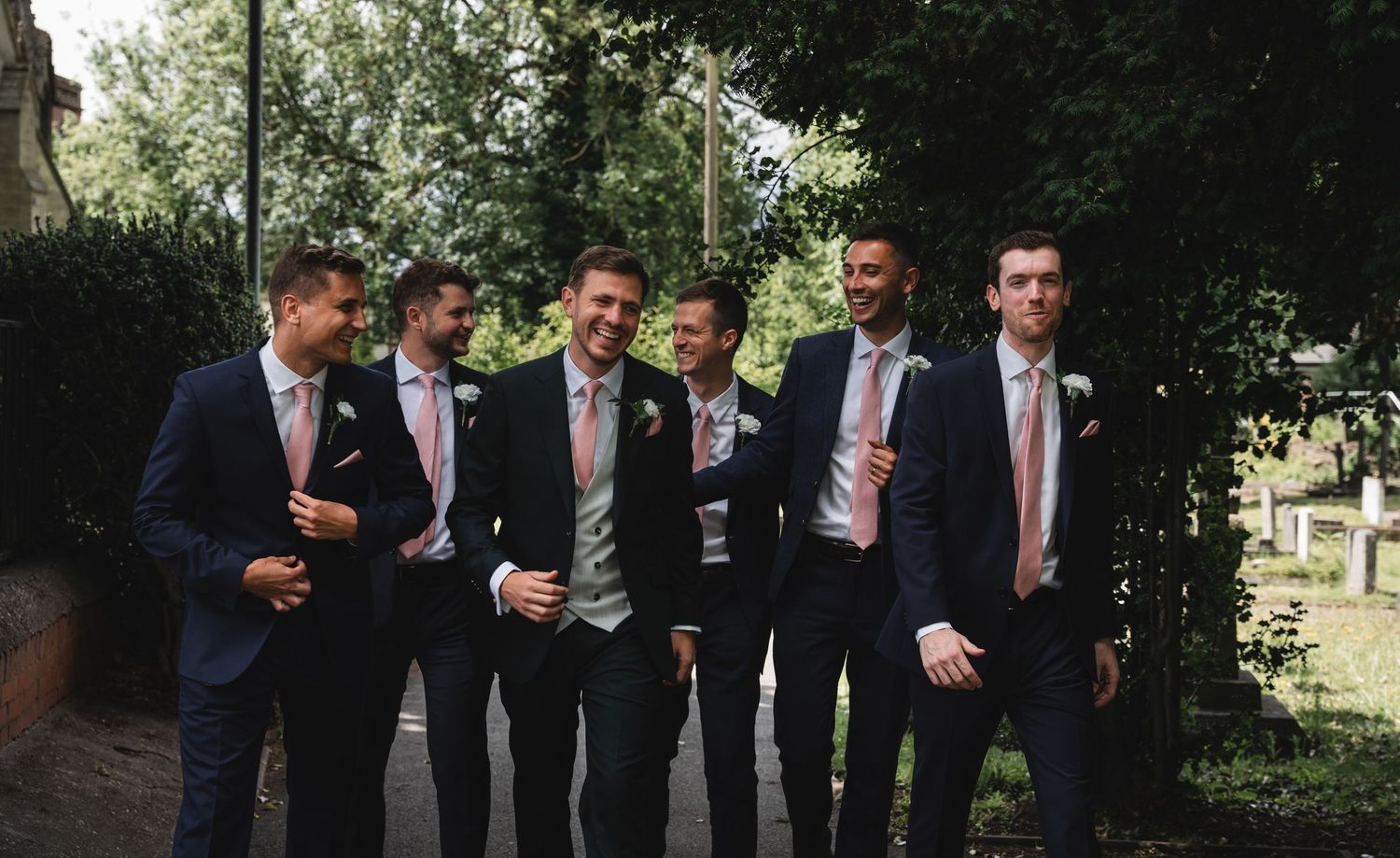 A group of groomsmen in navy suits and pink ties pose together outdoors for wedding photos.