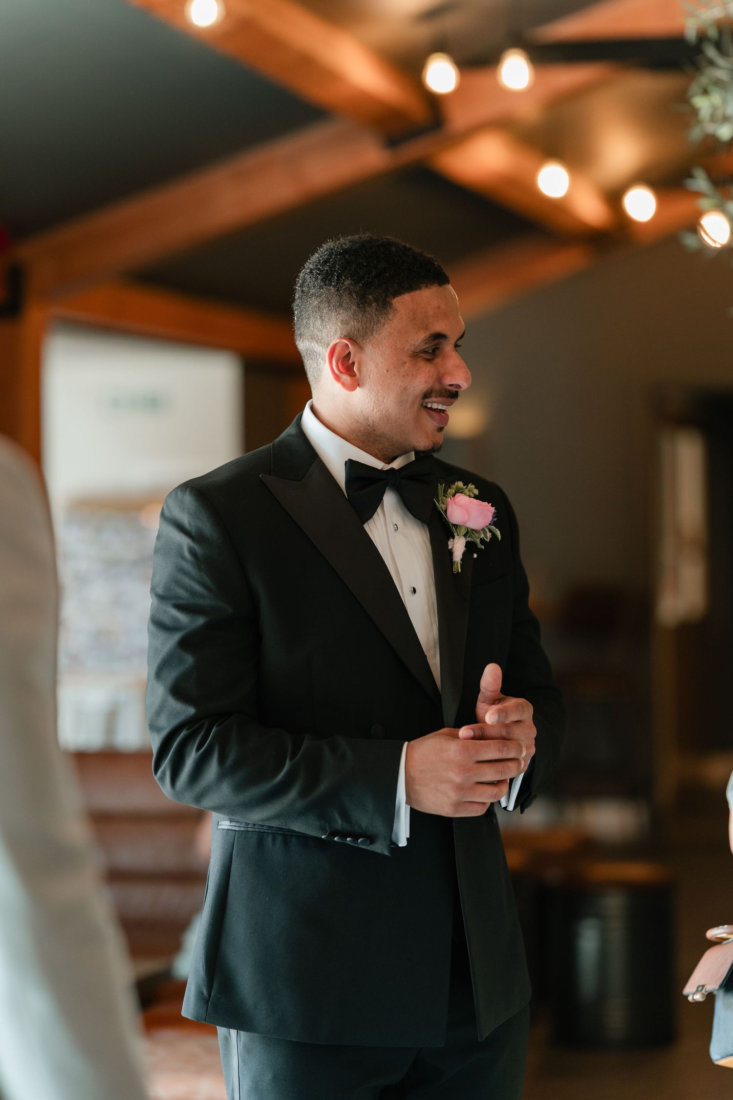 A groom in a classic black tuxedo with pink boutonniere laughs joyfully during an indoor wedding reception.