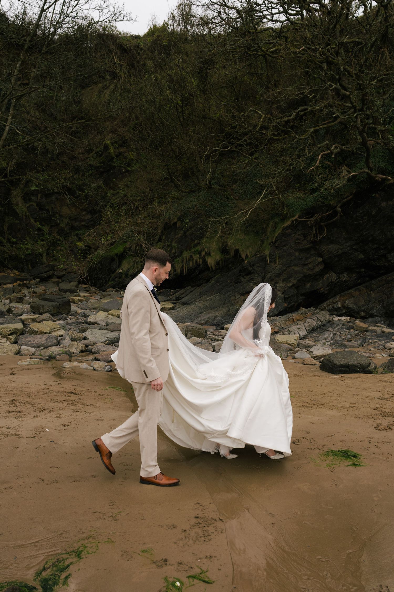 Groom in beige suit lifts bride's white gown as they walk on sandy beach with rocky cliffs behind.